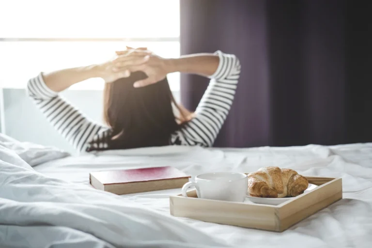 Une femme assise contre son lit où se trouve son petit-déjeuner