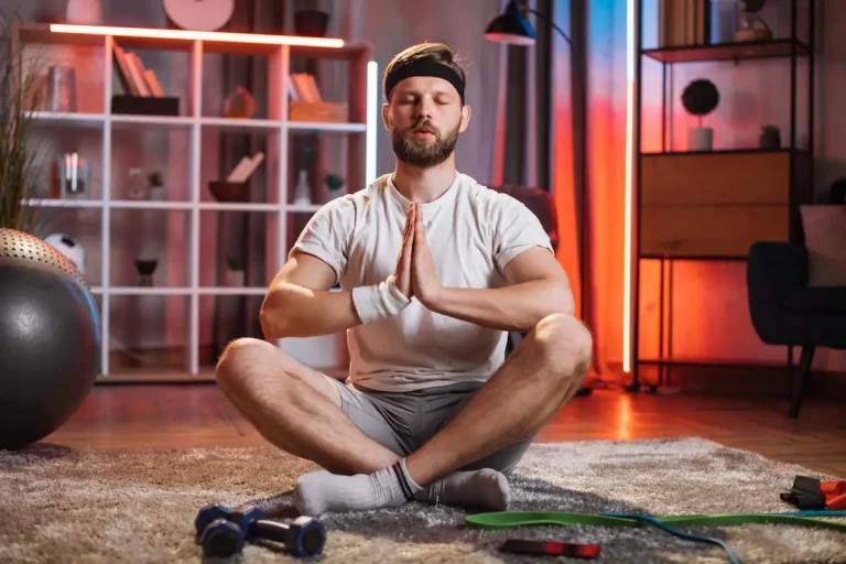 Un homme en position lotus au milieu du salon avec des équipements de sports autour de lui