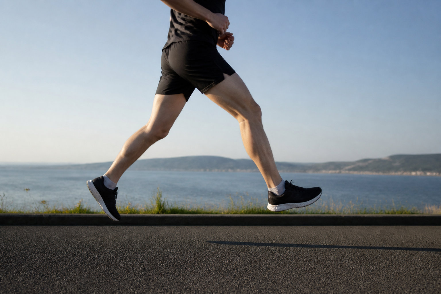 Un coureur en tenue noire court sur une route en bord de mer, focalisant sur le mouvement de ses jambes et du bassin.