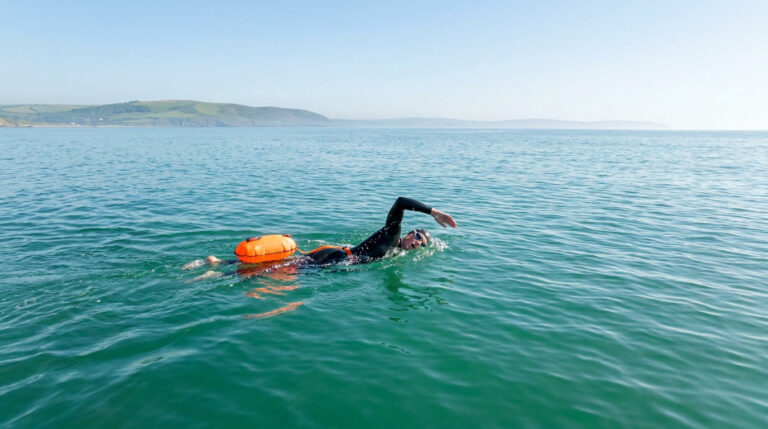 Un nageur en combinaison avec une bouée de sécurité orange nage dans une mer calme sous un ciel dégagé.